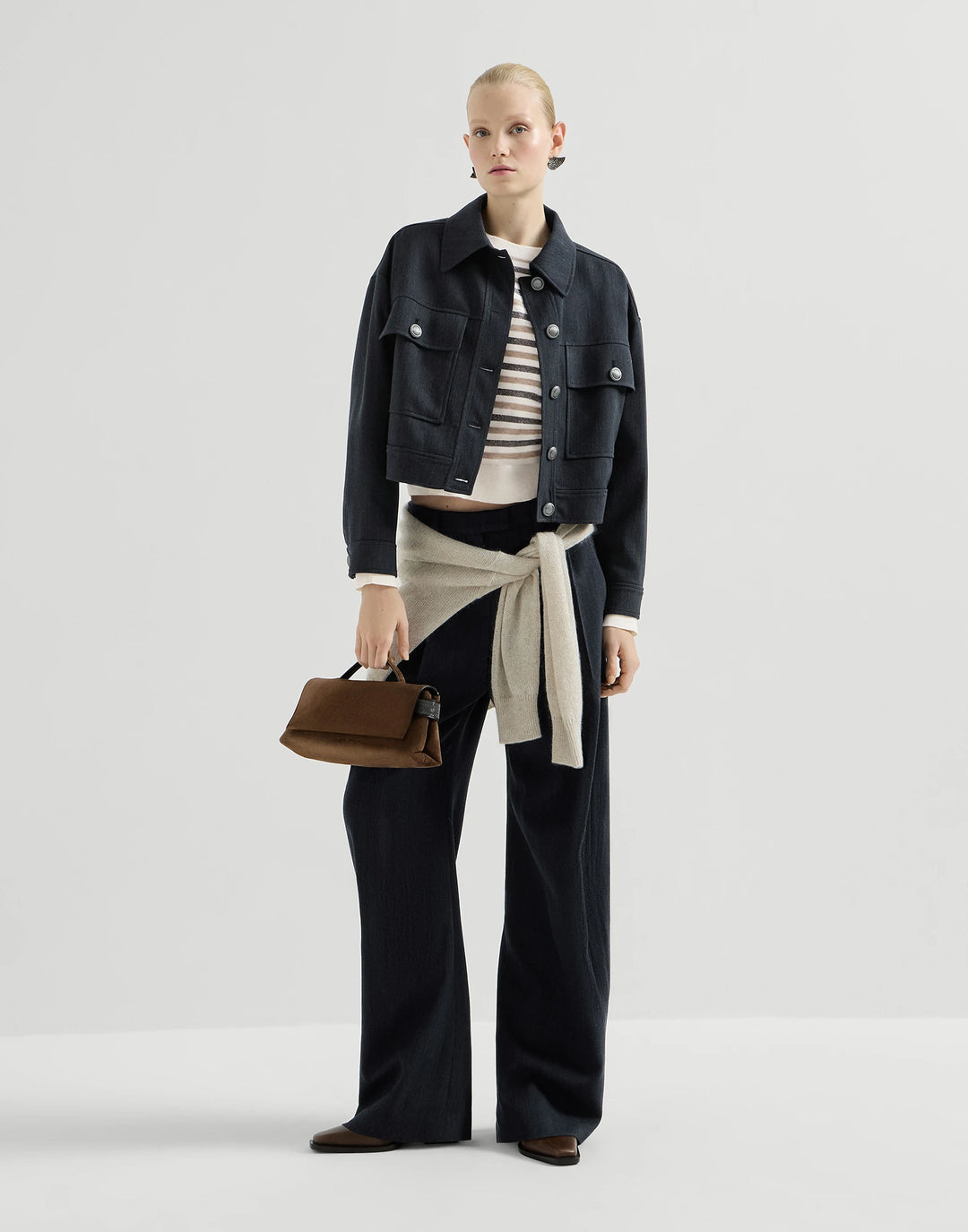 A model stands against a light gray studio background wearing a dark cropped denim jacket over a striped top, wide-leg dark pants, a beige tied sweater, and holding a brown leather handbag.