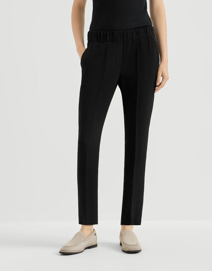 A model wearing black cropped trousers with an elastic waistband, paired with a black top and beige slip-on shoes, against a light gray studio background.
