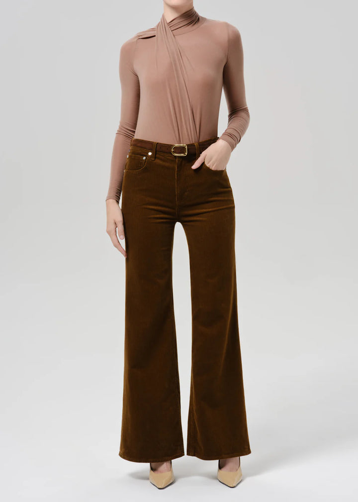 A model wearing a brown long-sleeve top with a unique draped neckline and dark brown wide-leg corduroy pants, styled with beige heels against a light gray studio background.