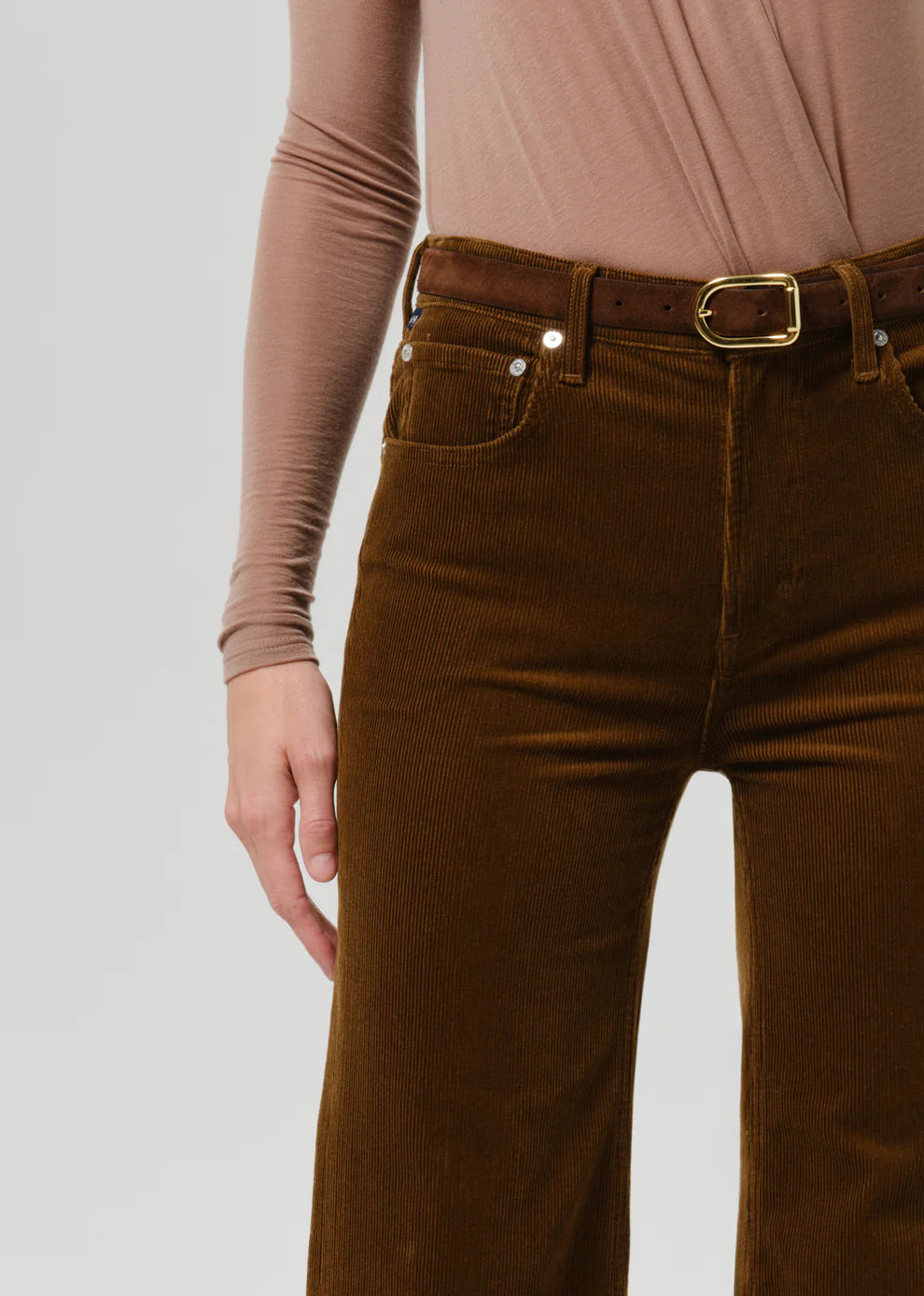 Close-up of a person wearing brown corduroy pants with a brown belt and gold buckle, paired with a dusty pink long-sleeve top, against a light gray studio background.