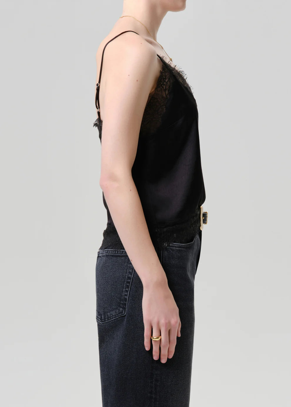 Side profile of a model wearing a black satin camisole top with dark lace trim and a gold-tone belt, paired with dark denim jeans against a light gray studio background.