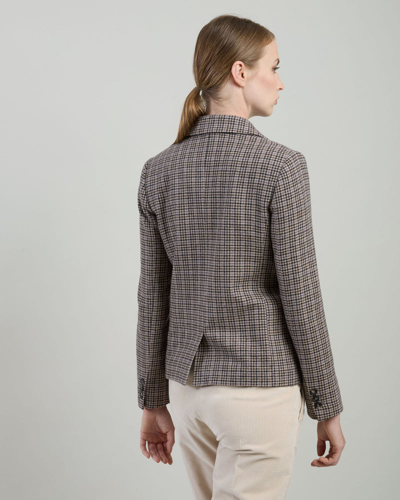 A woman shown from the back wearing a tailored blazer with a fine brown, beige, and black plaid pattern, paired with cream trousers, against a neutral light gray studio background.