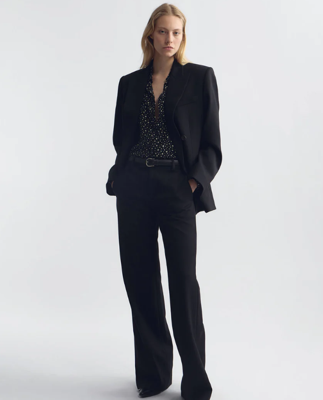 A model stands against a light gray studio background wearing a black tailored blazer and matching wide-leg trousers over a black embellished top.
