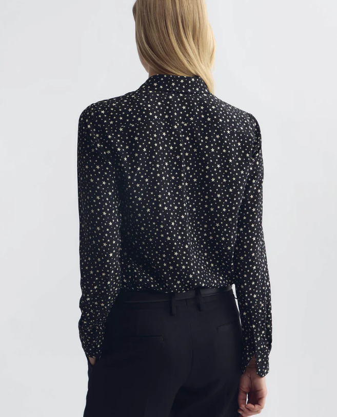 A woman shown from the back wearing a black long-sleeve blouse with a small white polka dot pattern and black trousers, against a plain light gray studio background.