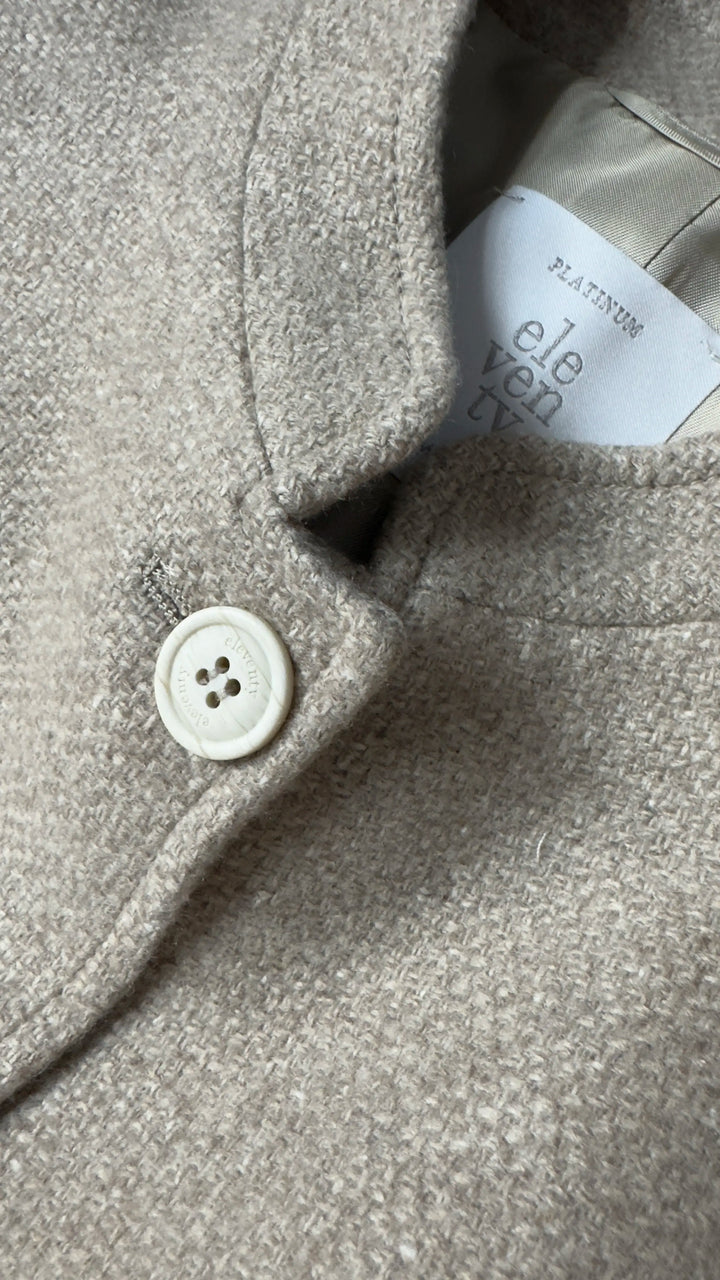 Close-up of a textured light beige wool coat with a white button and a visible interior label reading 'eleven ten'.