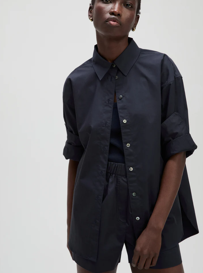 A model wearing a dark navy blue oversized button-down shirt with rolled-up sleeves and matching shorts, photographed against a plain light gray studio background.