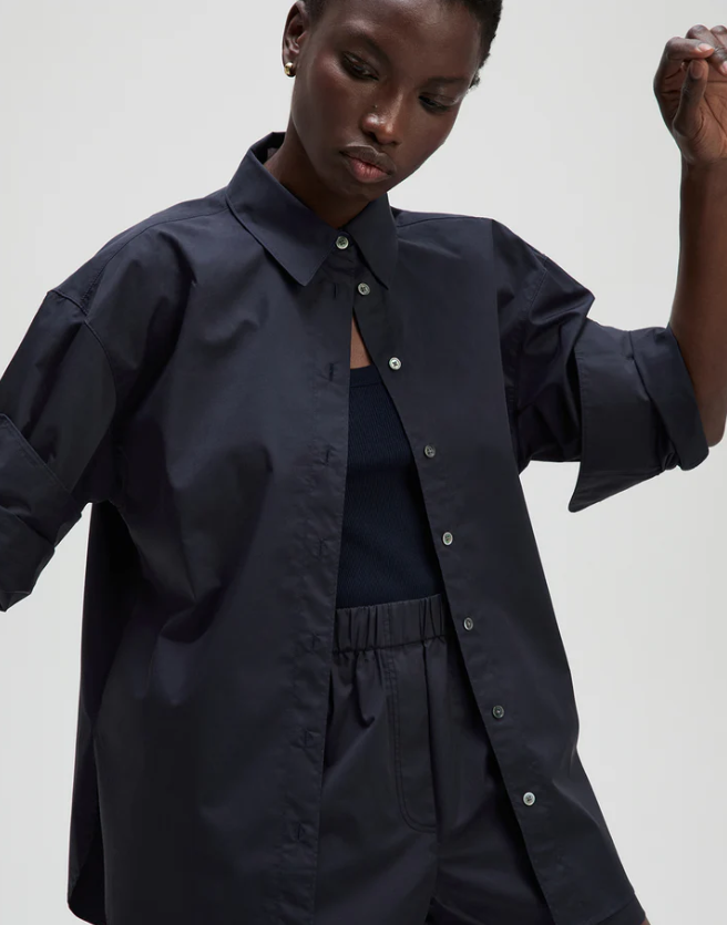 A model wearing an oversized dark navy blue button-up shirt with voluminous sleeves, styled over a matching dark top and trousers against a light gray studio background.