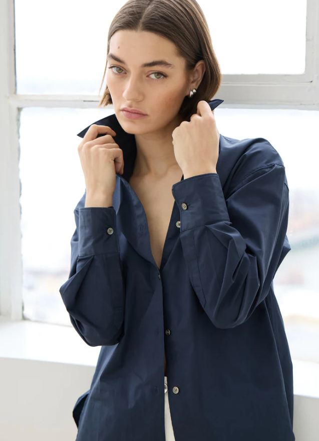 A woman wearing an oversized navy blue button-up shirt, standing in front of a bright window.