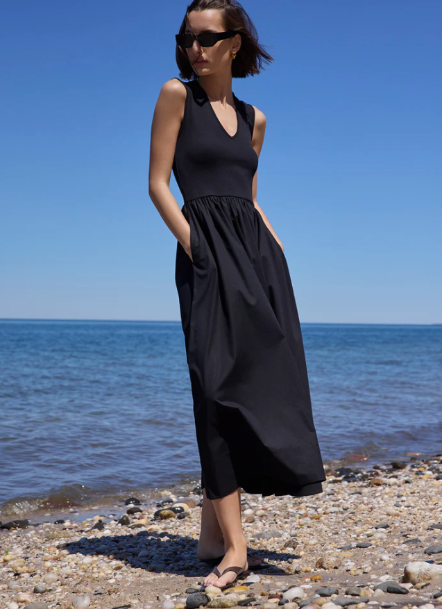 A woman wearing a black sleeveless maxi dress with side pockets stands on a pebble beach with the blue sea and sky in the background.