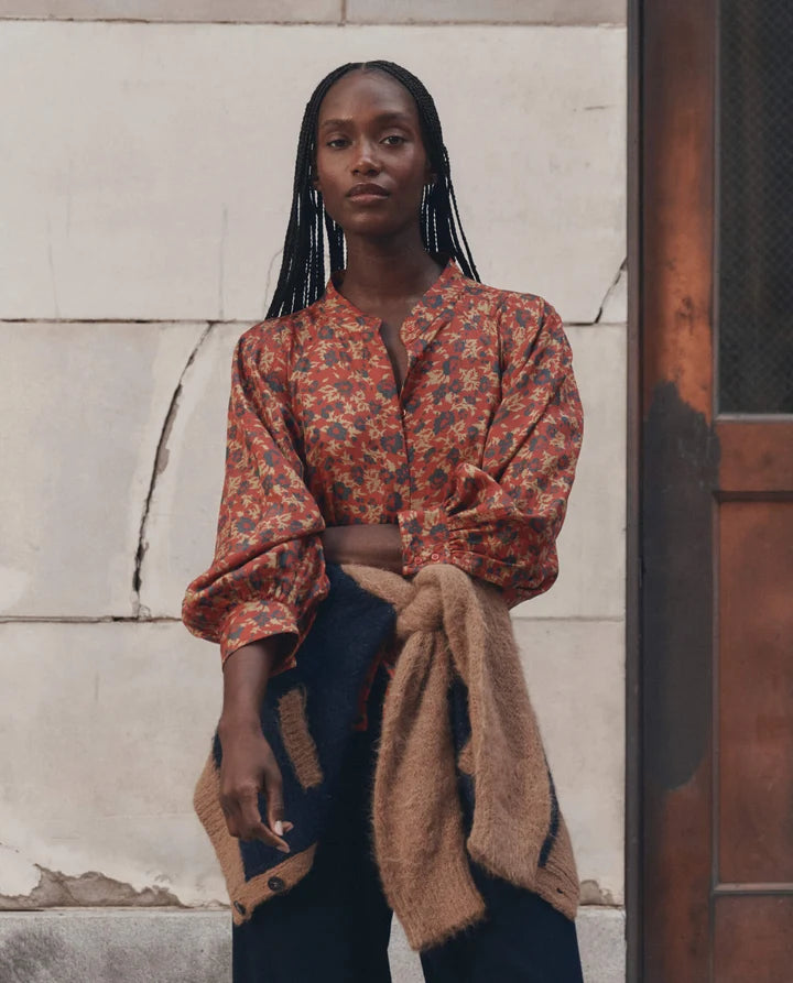 A woman stands outdoors against a light stone wall and dark wooden door, wearing a rust-orange floral long-sleeve blouse with dark blue pants and a tan knit wrap tied around her waist.