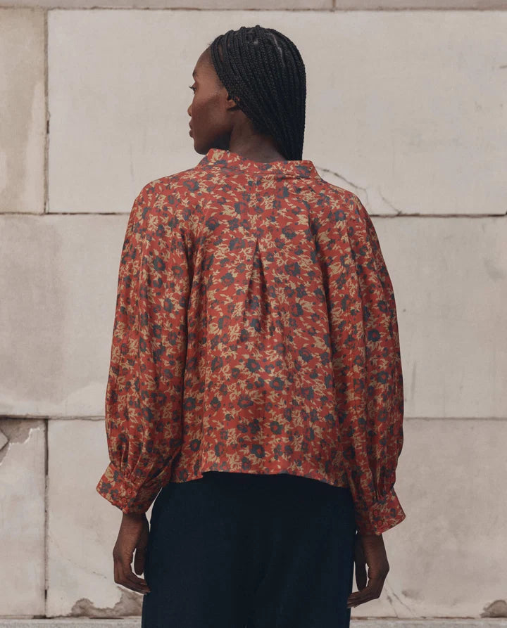 A woman viewed from behind wearing a long-sleeve blouse with a dense floral pattern in rust red, beige, and dark blue, paired with dark navy trousers, standing against a light stone wall.