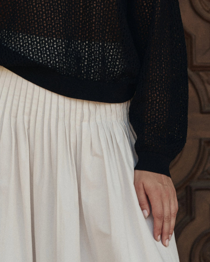 Person wearing a black sweater with a textured pattern and a white pleated skirt, standing against a wooden panel background.