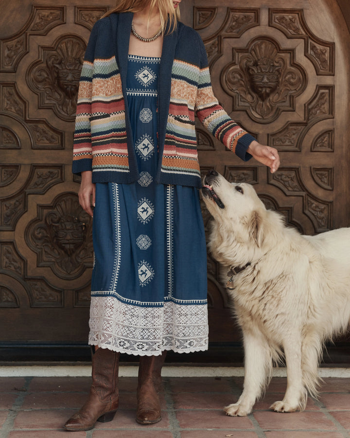 Person wearing a patterned cardigan and dress standing next to a white dog in front of a decorative wooden door.
