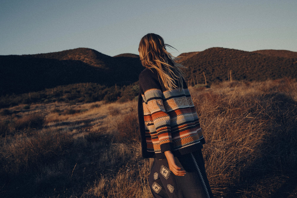Person wearing a patterned sweater standing in a desert landscape with mountains in the background