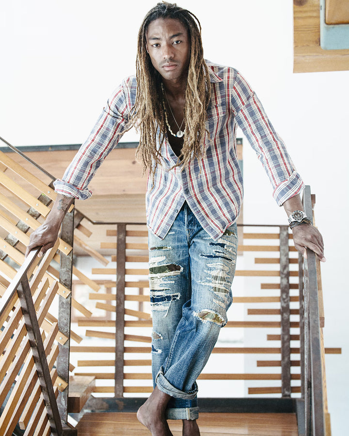 Man wearing a plaid shirt and ripped jeans standing on a wooden staircase.
