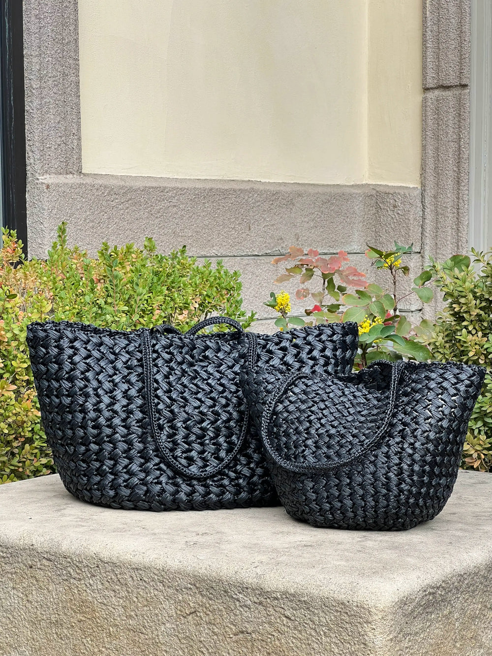 Two black woven handbags on a stone ledge with a garden background