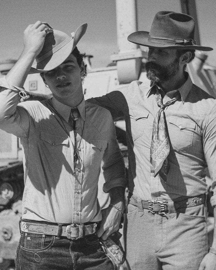 Two men in cowboy attire standing outdoors with a vehicle in the background.
