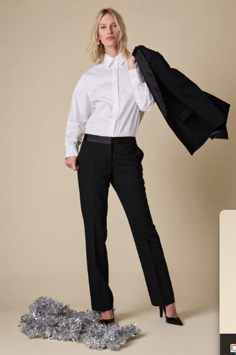 Woman in a white blouse and black pants holding a black coat against a beige background