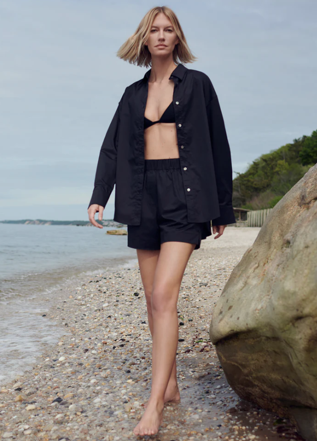 Woman in black outfit walking on a beach with a large rock and water in the background