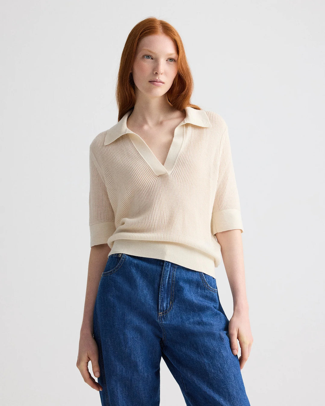 Woman wearing a beige knit polo shirt and blue jeans on a light gray background
