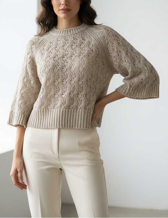 Woman wearing a beige knitted sweater and white pants against a plain background