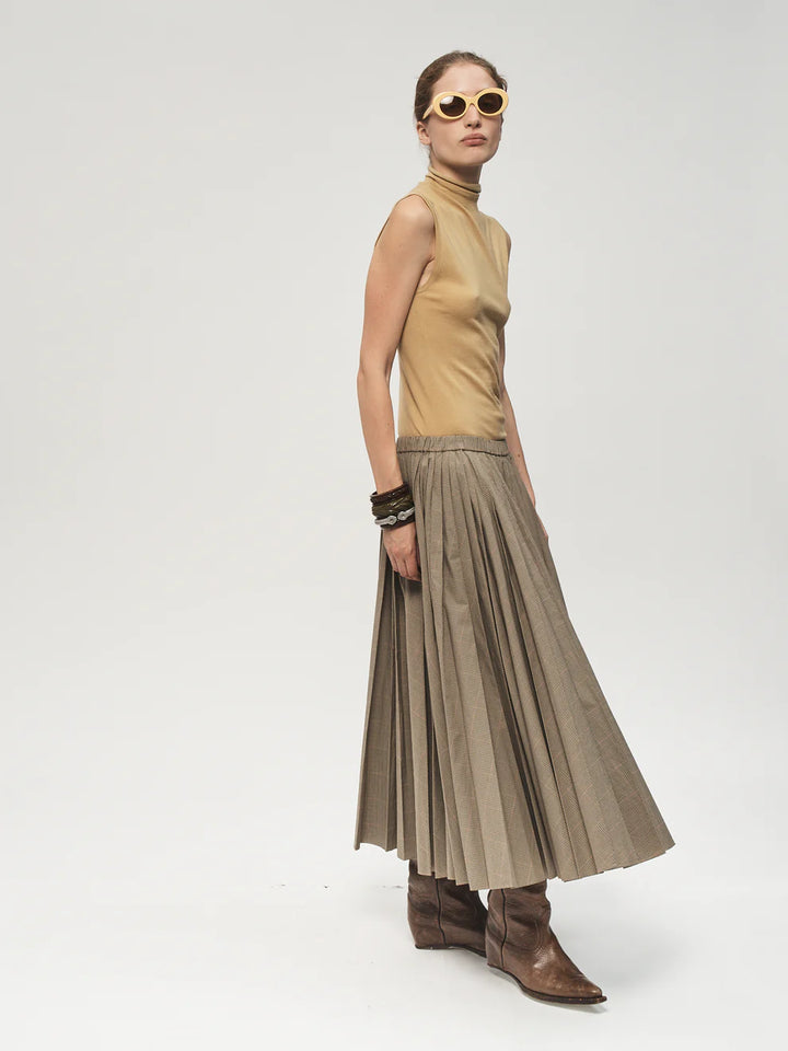 Woman wearing a beige sleeveless top and pleated skirt with brown boots on a white background
