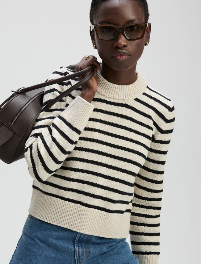 Woman wearing a black and white striped sweater holding a brown handbag against a light gray background