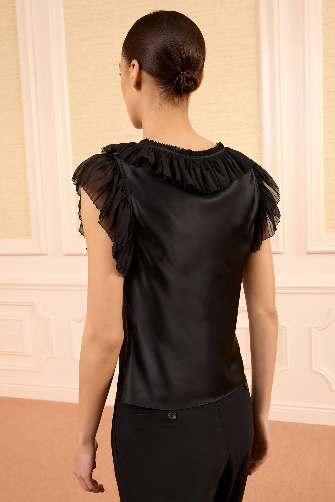 Woman wearing a black blouse with ruffled shoulders against a beige wall.