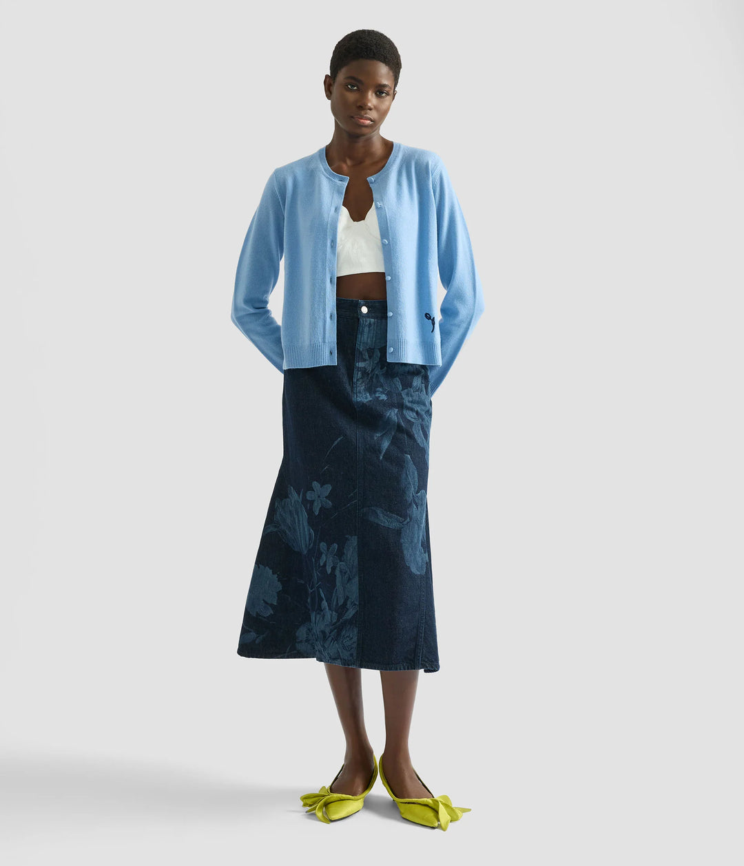 Woman wearing a blue cardigan, white top, and dark blue skirt on a light gray background