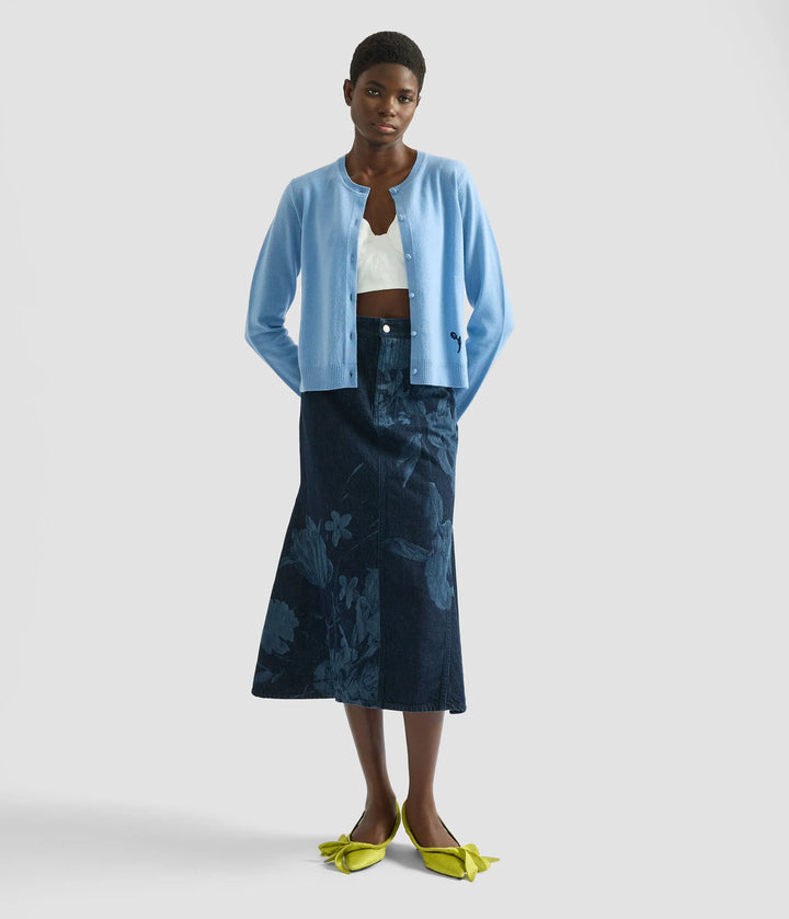 Woman wearing a blue cardigan, white top, and dark blue skirt on a light gray background