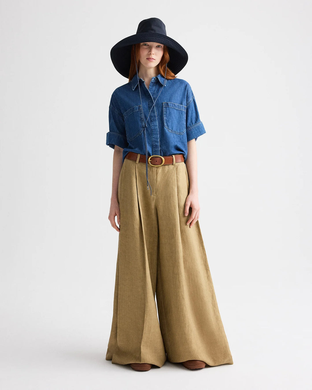 Woman wearing a blue denim shirt, wide-leg beige pants, and a black hat on a white background