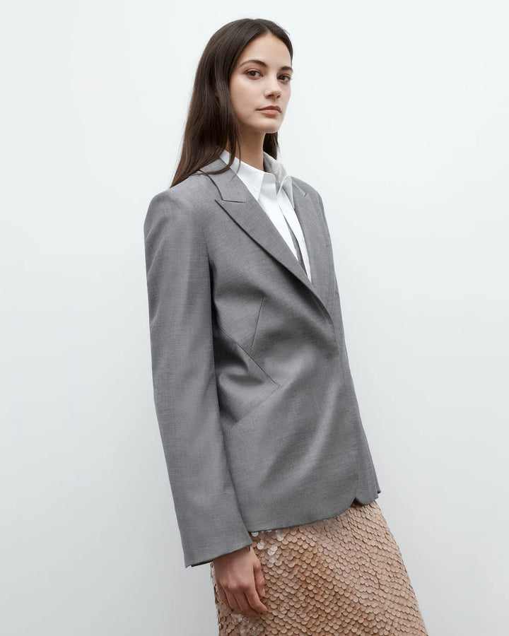 Woman wearing a gray blazer over a white shirt with a beige skirt on a plain background