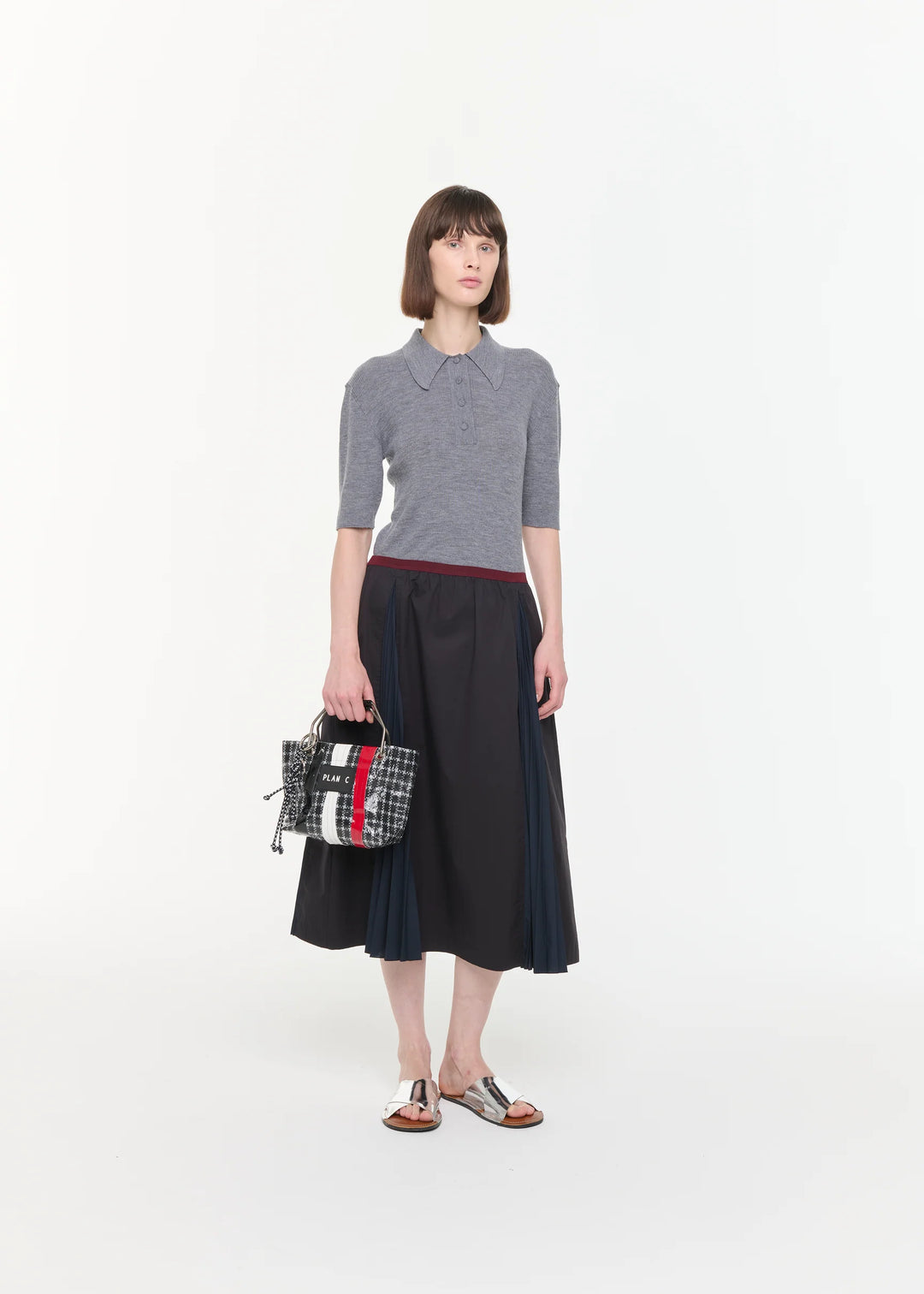 Woman wearing a gray sweater and dark skirt holding a patterned handbag on a white background