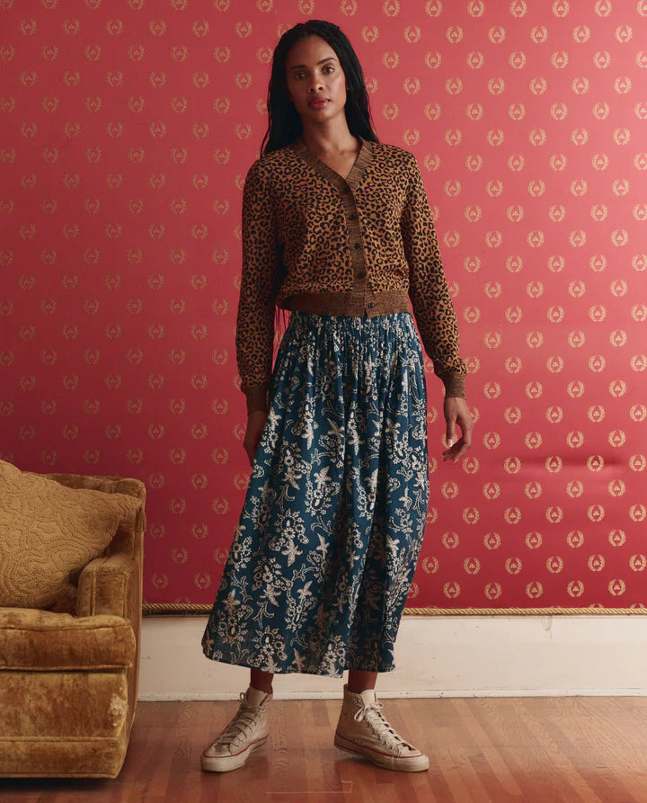 Woman wearing a leopard print top and blue floral skirt standing in a room with patterned wallpaper.