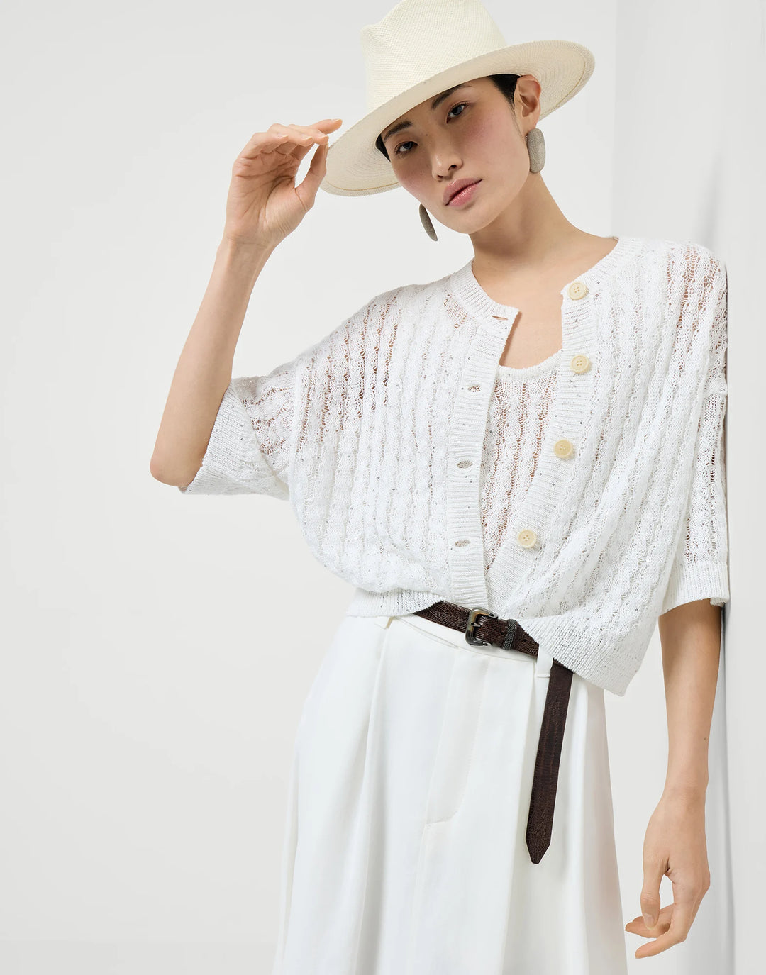 Woman wearing a white knit cardigan over a white dress with a wide-brimmed hat against a plain background