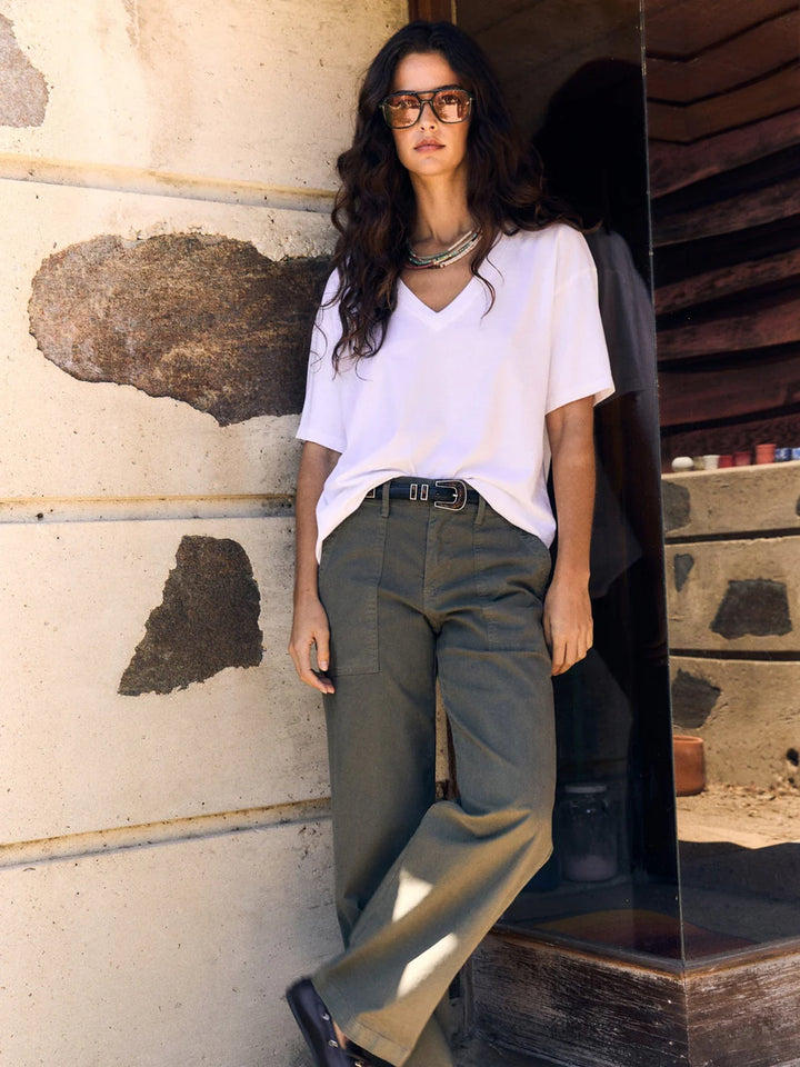 Woman wearing a white shirt and green pants standing against a textured wall.