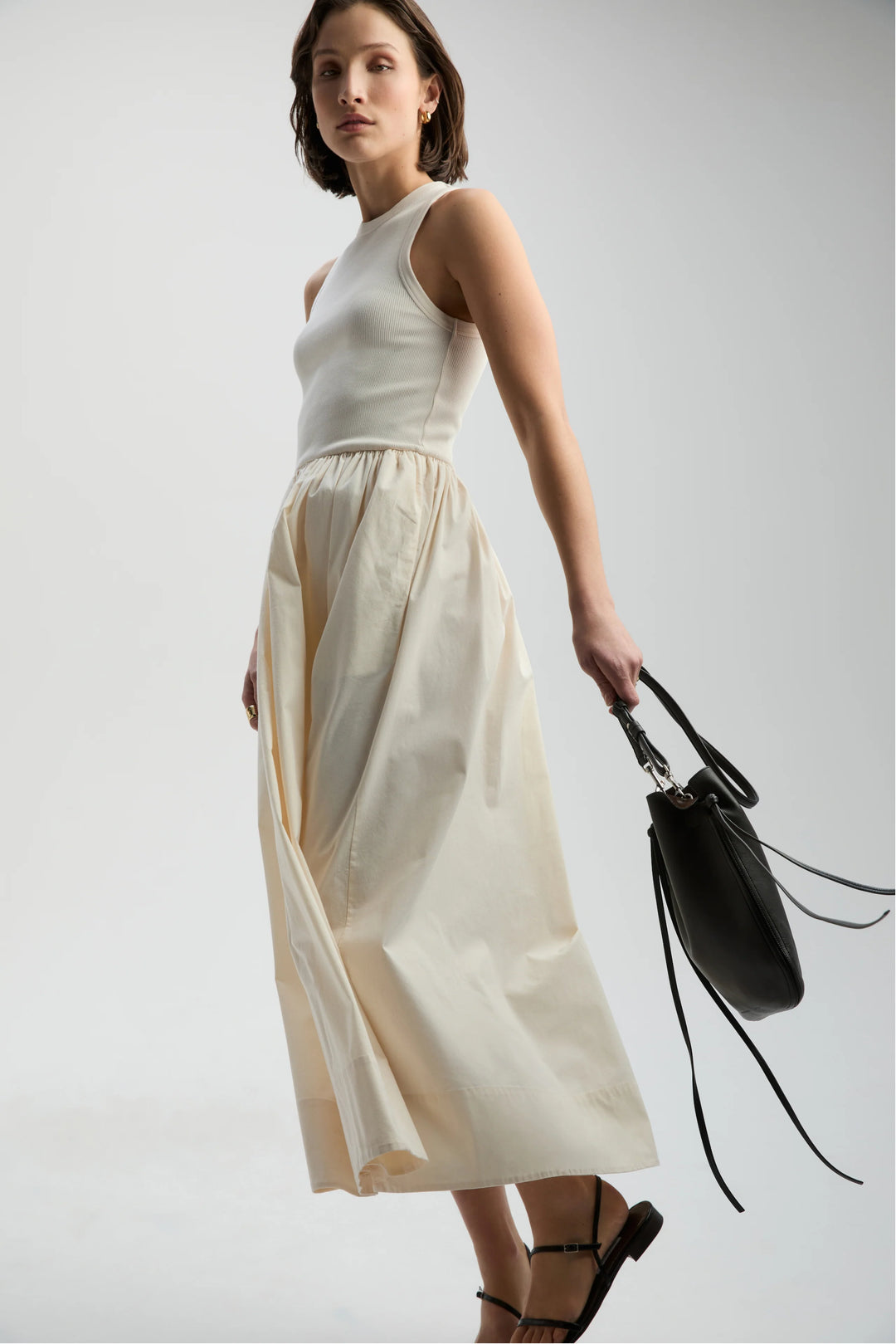 Woman wearing a white sleeveless top and beige skirt holding a black handbag on a light gray background