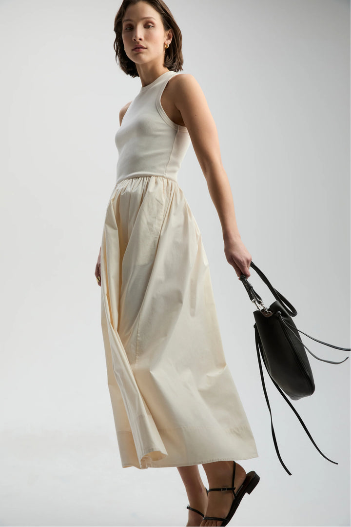 Woman wearing a white sleeveless top and beige skirt holding a black handbag on a light gray background