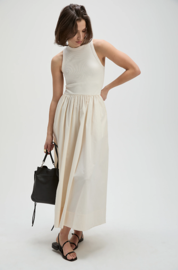 Woman wearing a white sleeveless top and beige skirt holding a black handbag on a white background