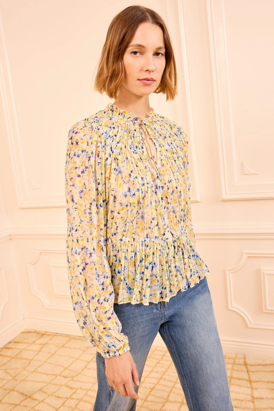 Woman wearing a yellow floral blouse and blue jeans against a beige wall.