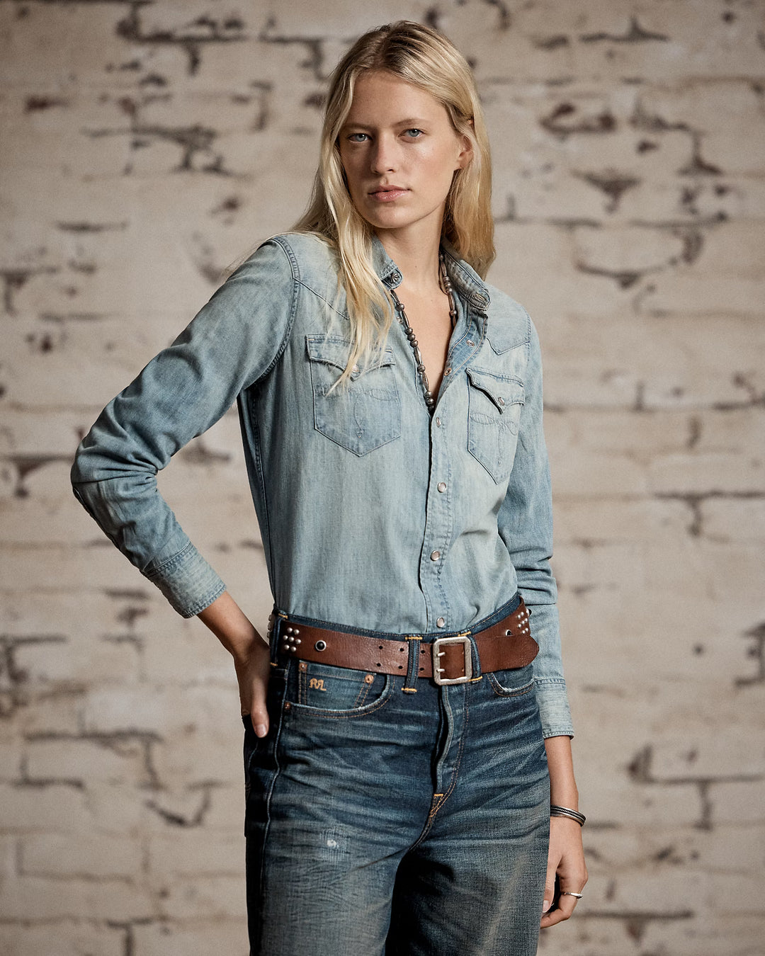 Woman wearing a denim shirt and jeans against a brick wall.