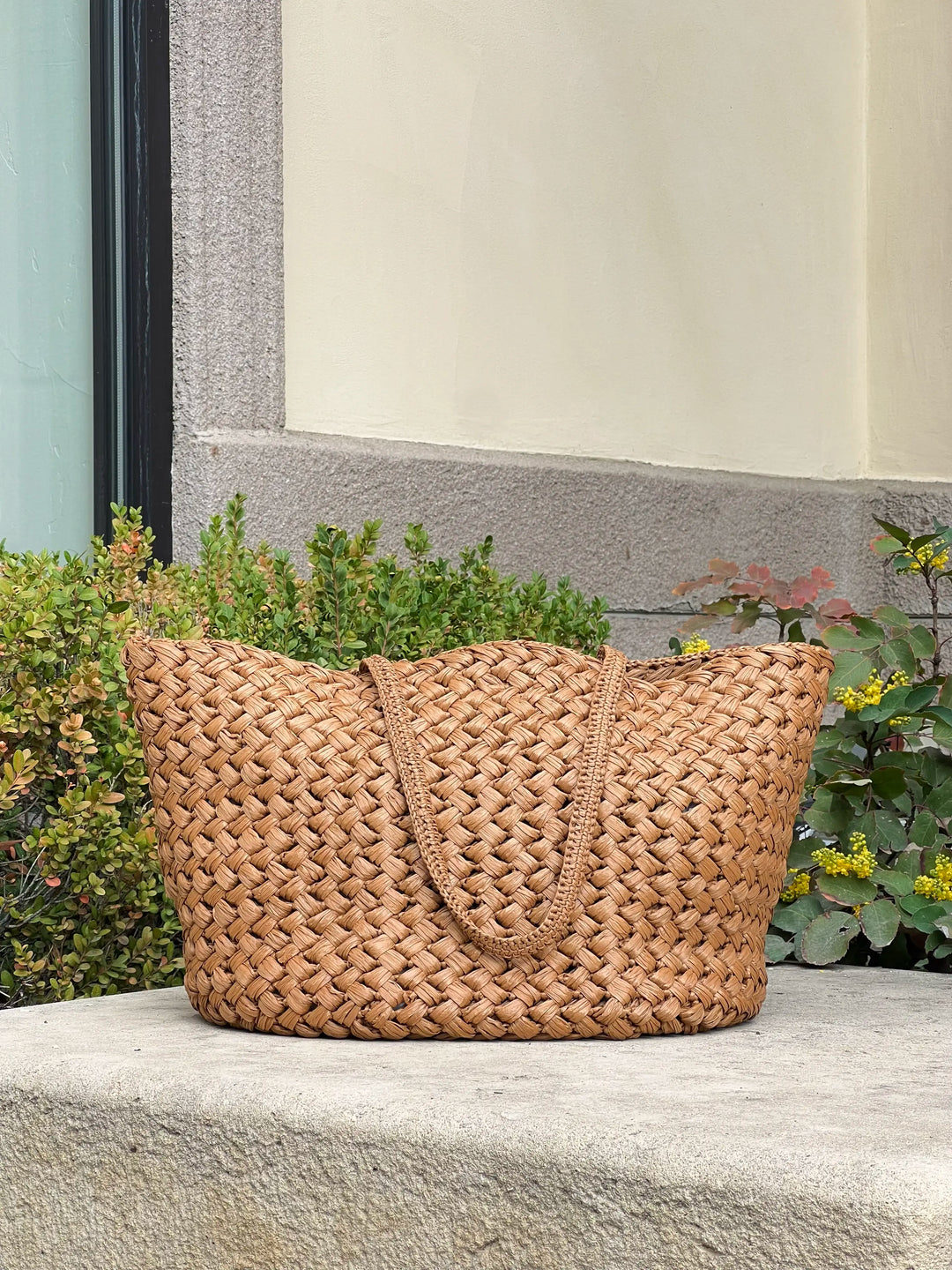 Woven brown bag on a stone surface with plants in the background