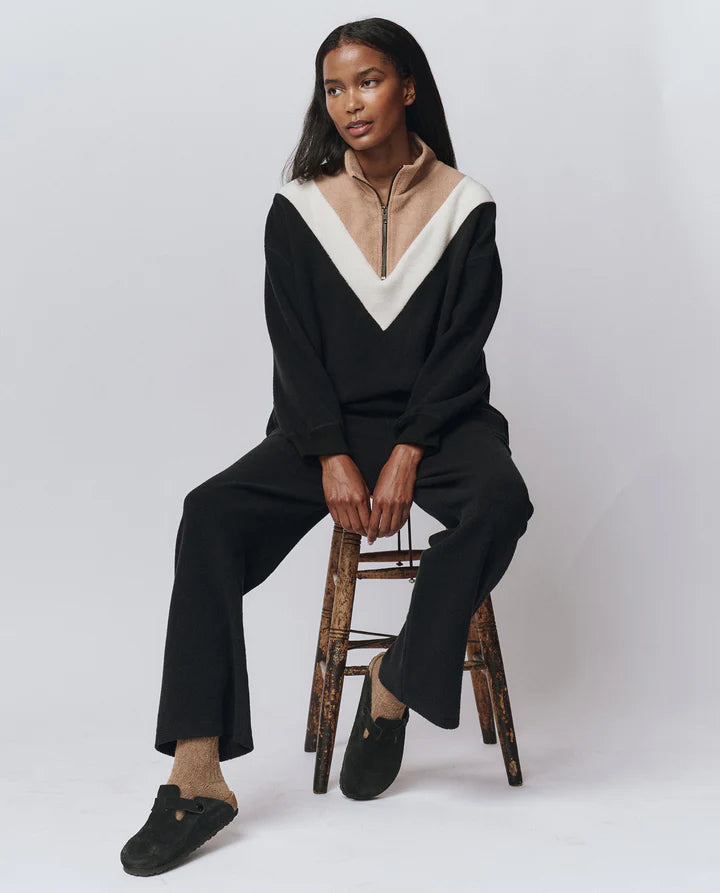 Person wearing a black and beige outfit sitting on a wooden stool against a white background