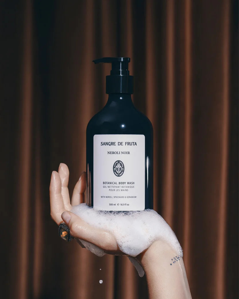 Hand holding a bottle of 'Sangre de Fruta' body wash with a brown curtain background