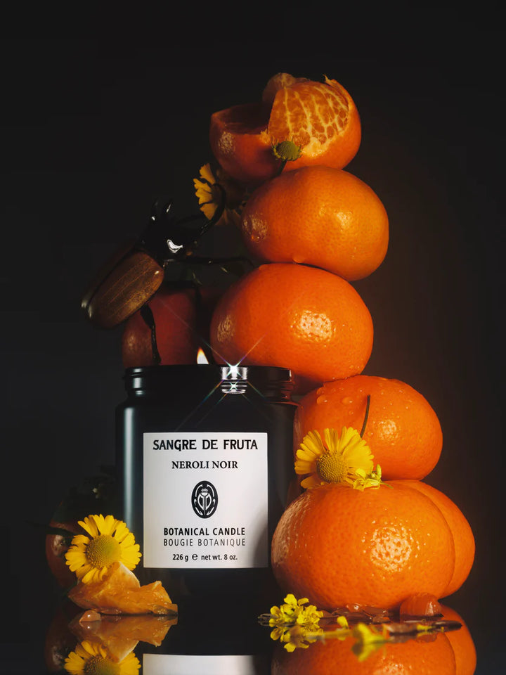 Sangre de Fruta Neroli Noir candle surrounded by oranges and flowers on a dark background