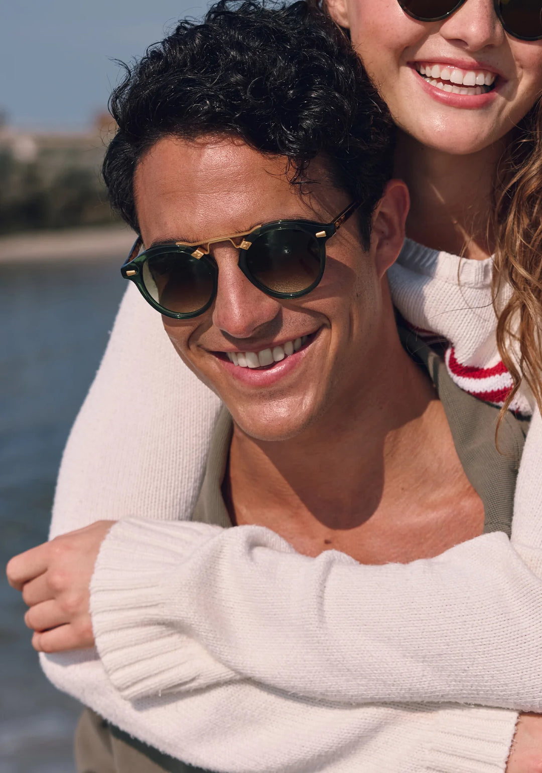 Man and woman smiling at the camera with sunglasses on, standing by a body of water.
