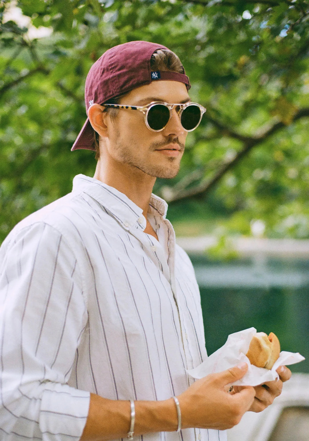 Man wearing sunglasses and a maroon cap, holding a hot dog outdoors.