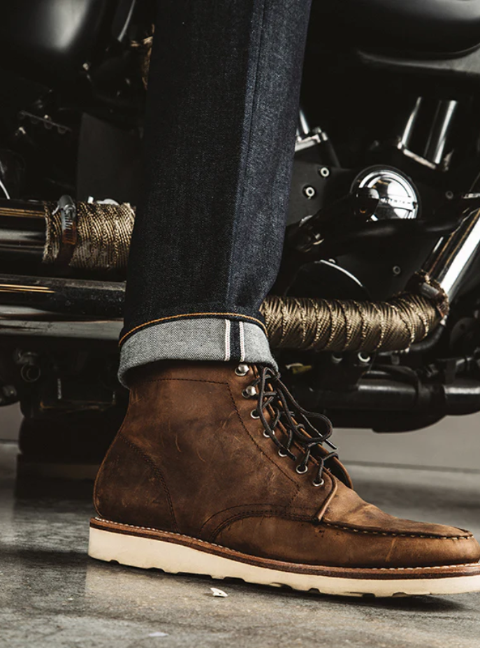 Brown leather boot worn with rolled-up jeans against a motorcycle background