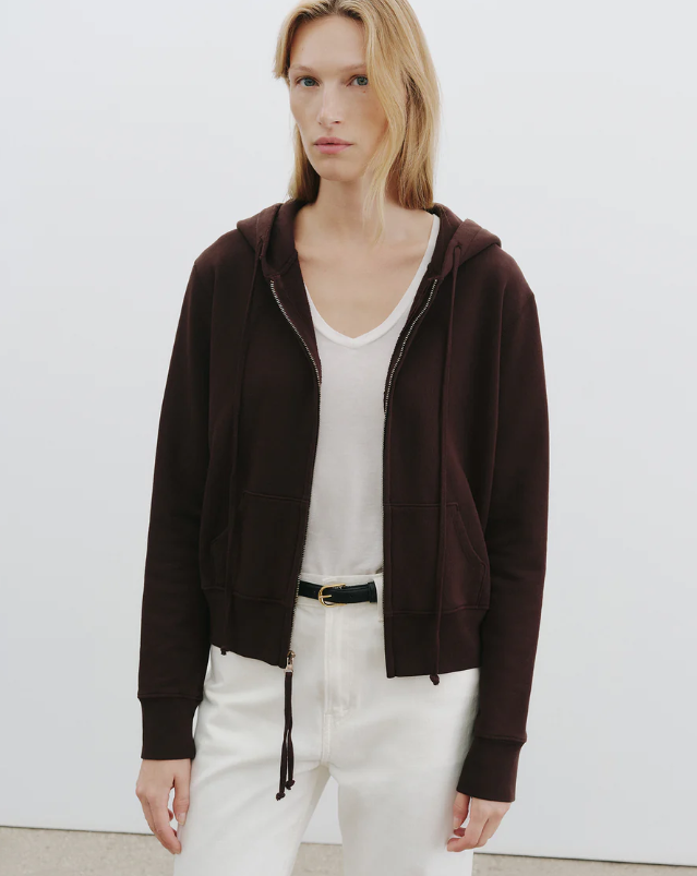 Person wearing a brown zip-up hoodie over a white shirt and pants on a plain background