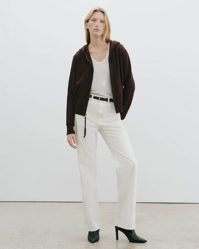 Woman wearing a brown jacket, white top, and white pants on a plain background